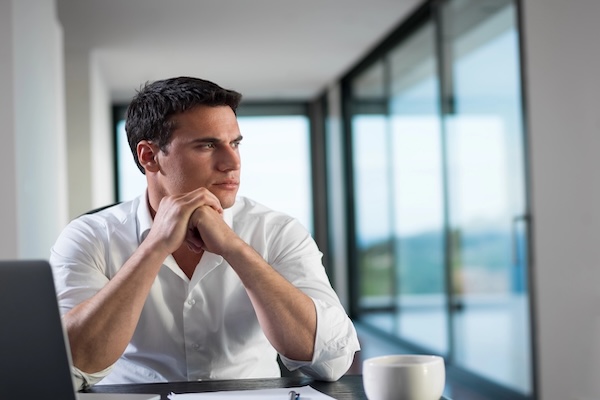 Man sitting at a desk, looking out a window thoughtfully while working on a laptop in a modern office