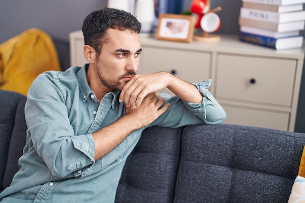 Man sitting on a couch at home, looking off to the side with a thoughtful, preoccupied expression