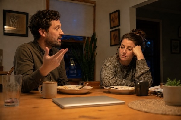 Man talking at dinner while his partner listens quietly and looks emotionally fatigued in a softly lit home kitchen
